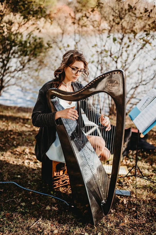 Alycen playing her black harp beneath trees beside the water